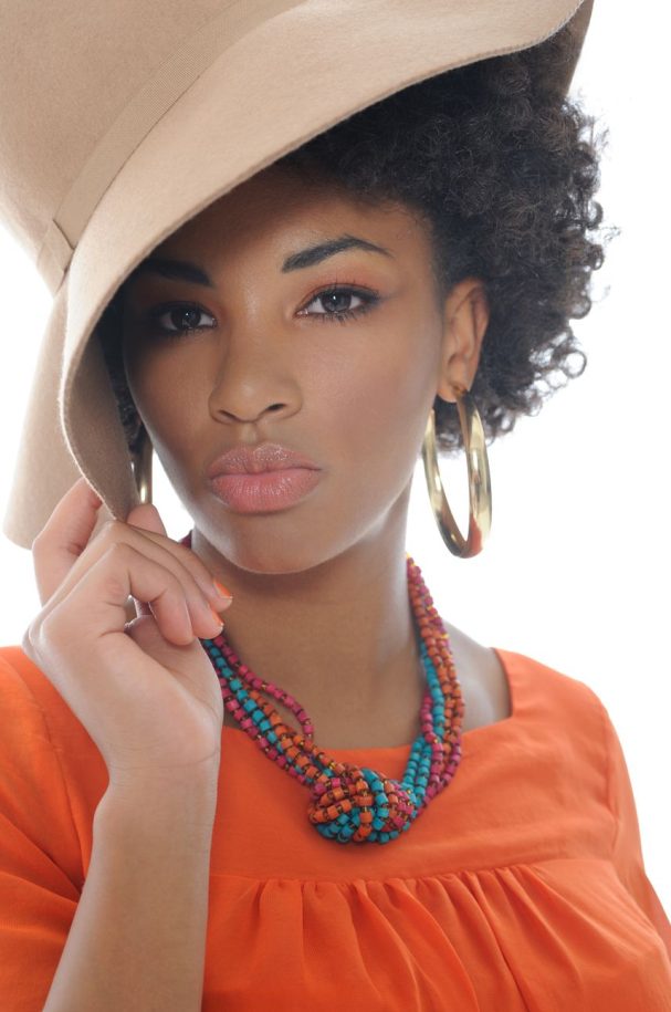 sweet every day Make-up Frau mit lockigem Haar, großem Hut und buntem Schmuck in orangefarbenem Shirt.