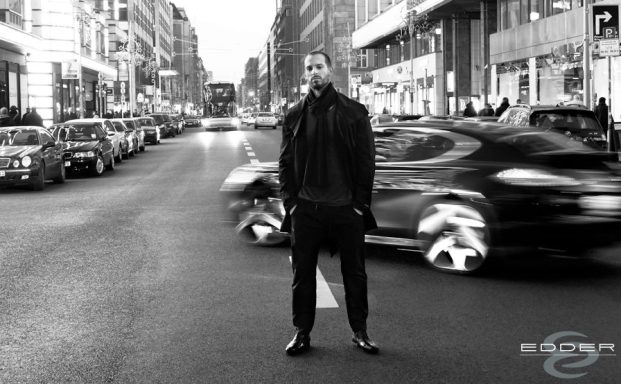Urban fashion by umasan Mann in schwarzer Jacke steht auf einer Straße, umgeben von Verkehr und Gebäuden.