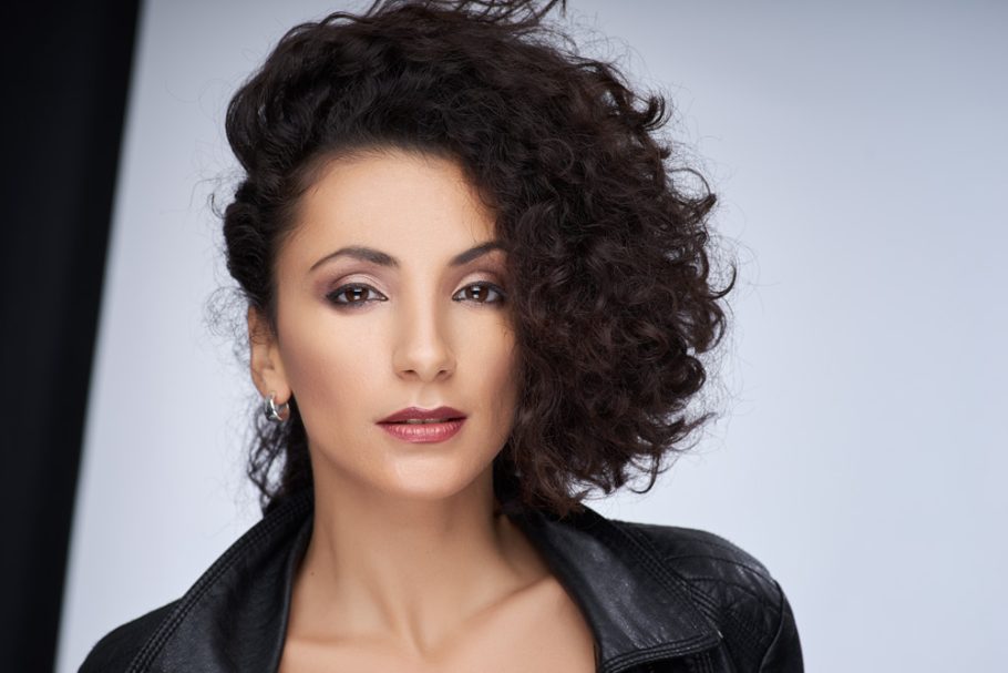 Business und Make-up Frau mit lockigem, braunem Haar und einem selbstbewussten Gesichtsausdruck.